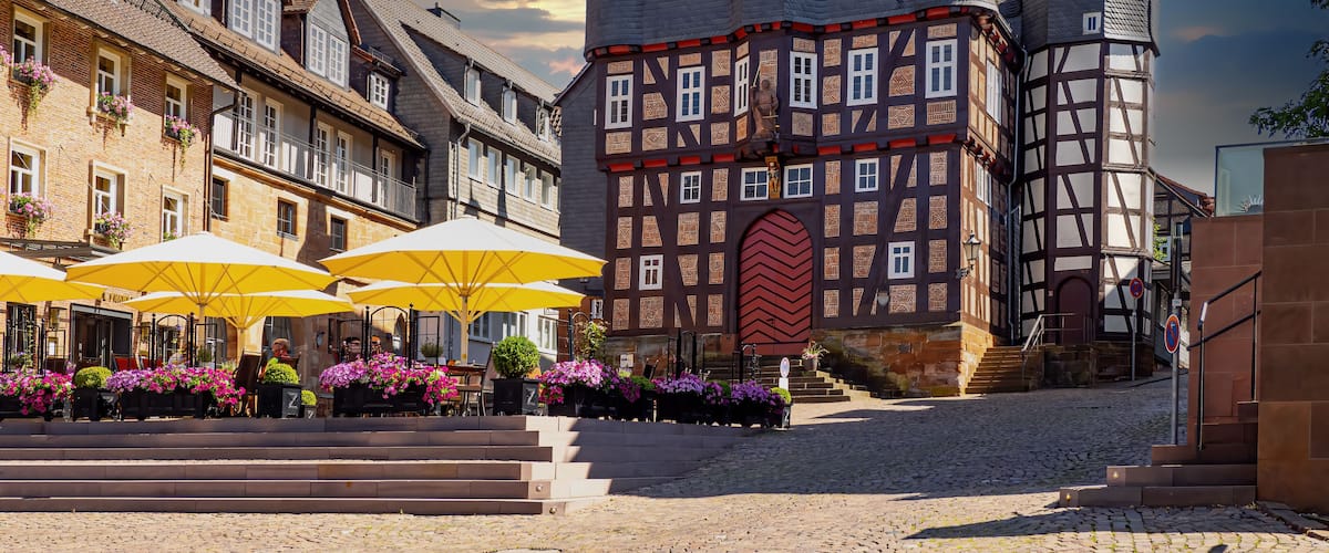 A historic European town square with traditional half-timbered buildings, a prominent clock tower, and outdoor seating with umbrellas.