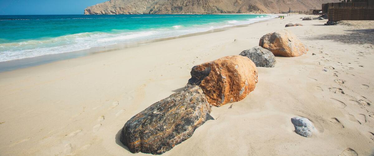 A sandy beach with large rocks in the foreground and a mountain range in the background.