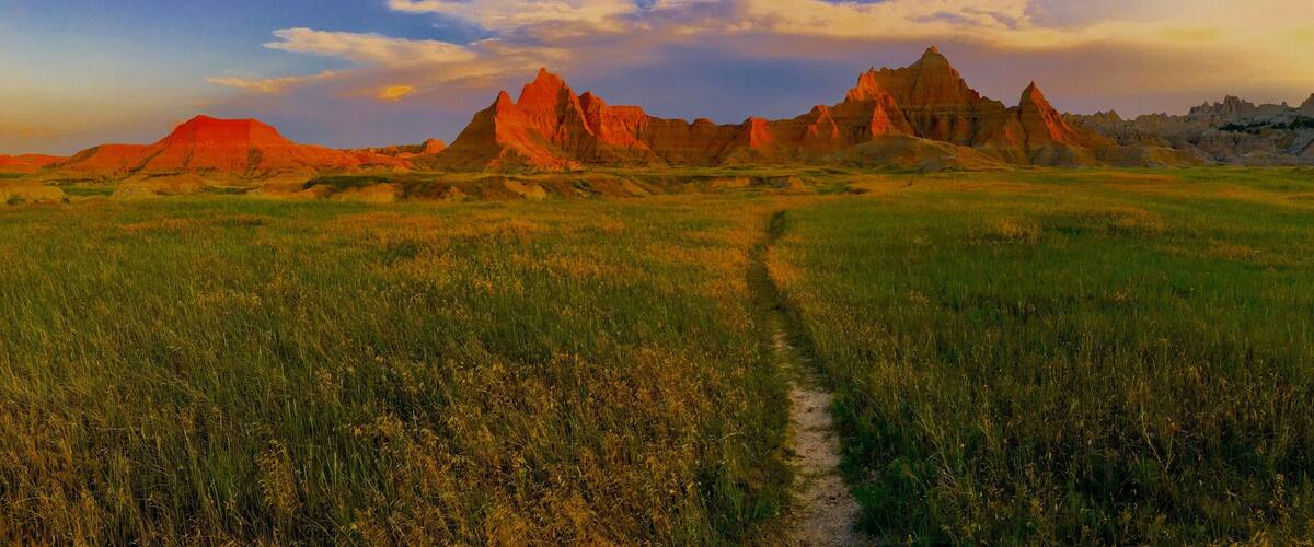 A field with a dirt path leading to a range of red rock formations.