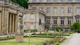 A historic building with classical architecture, a statue, and a well-maintained garden.