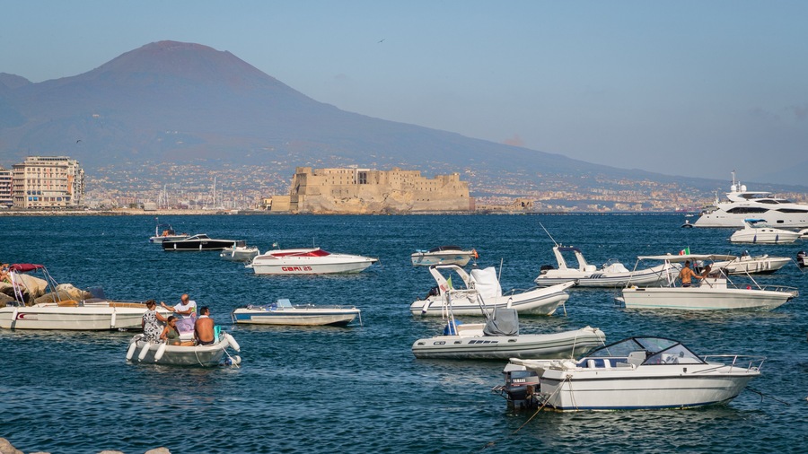 Un port de plaisance avec différents types de bateaux, un château imposant et une montagne en arrière-plan.