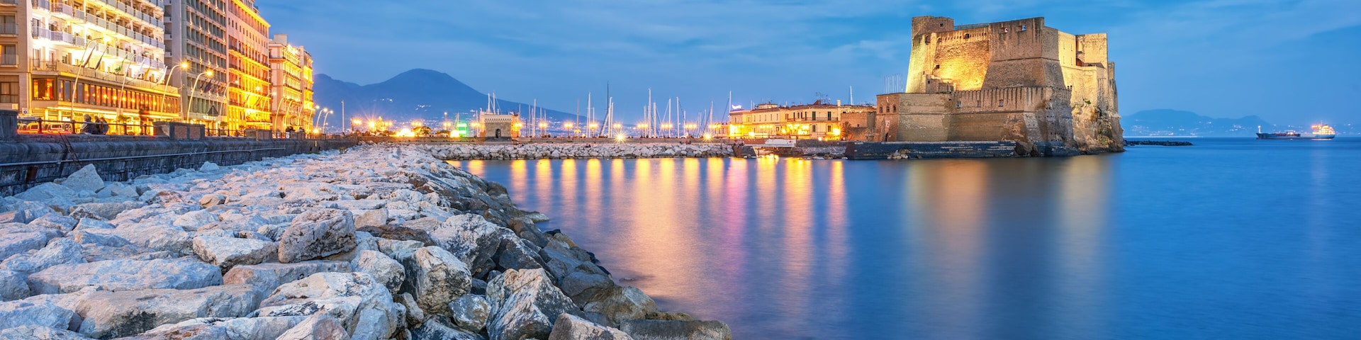Paisagem costeira com um castelo histórico, edifícios modernos e mar calmo.