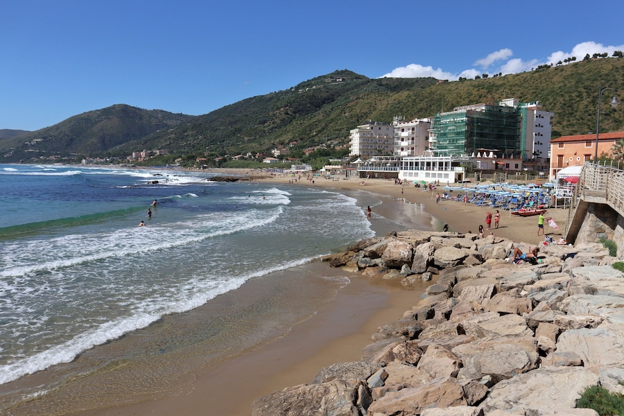 Una spiaggia con costa rocciosa, onde e montagne sullo sfondo.