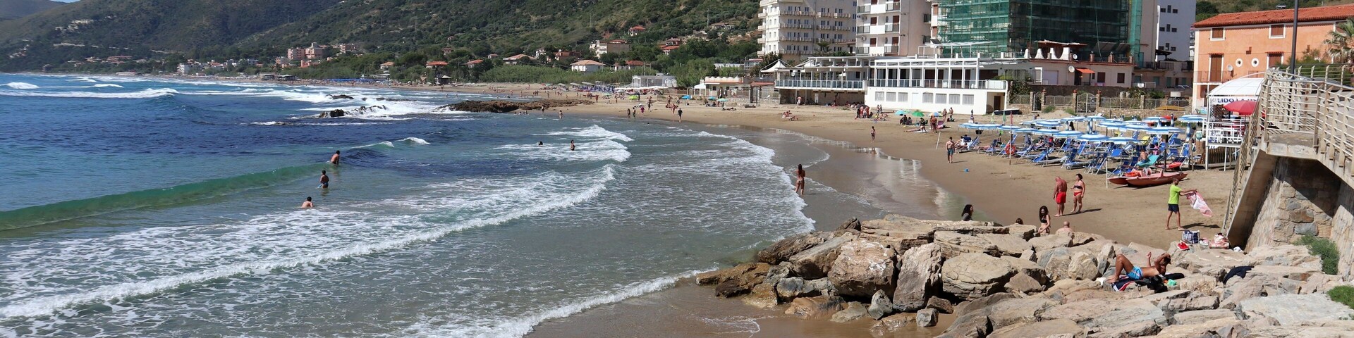 Una playa con costa rocosa, olas y montañas al fondo.