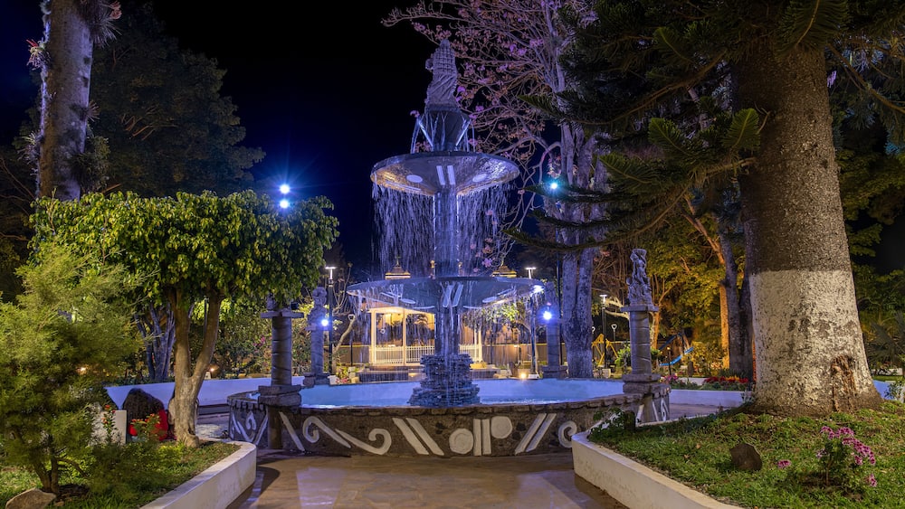 Une fontaine illuminée, avec des jets d’eau, entourée d’arbres et éclairée par des jeux de lumière.