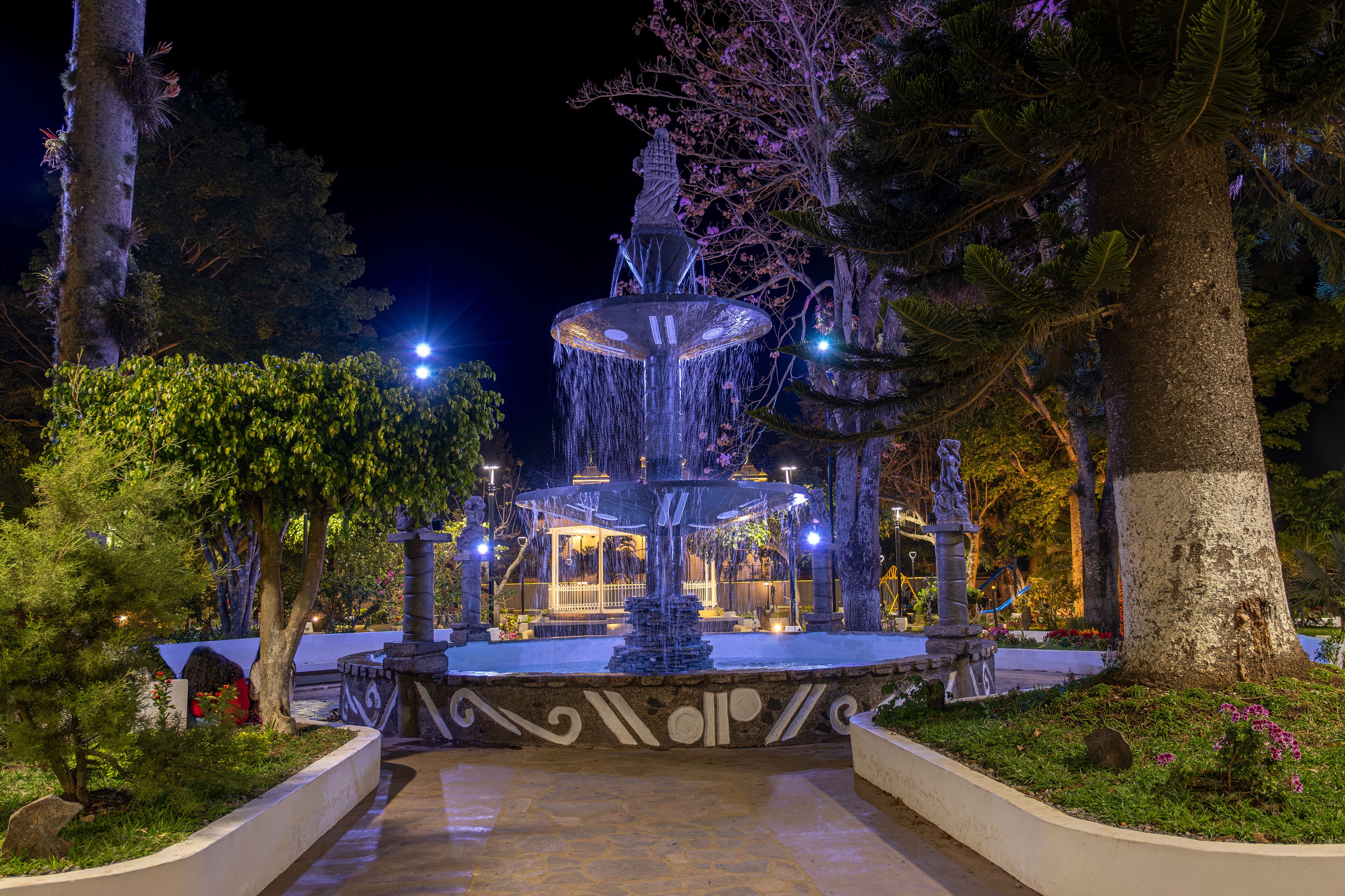 A nighttime fountain with water jets, surrounded by trees and illuminated by lights.