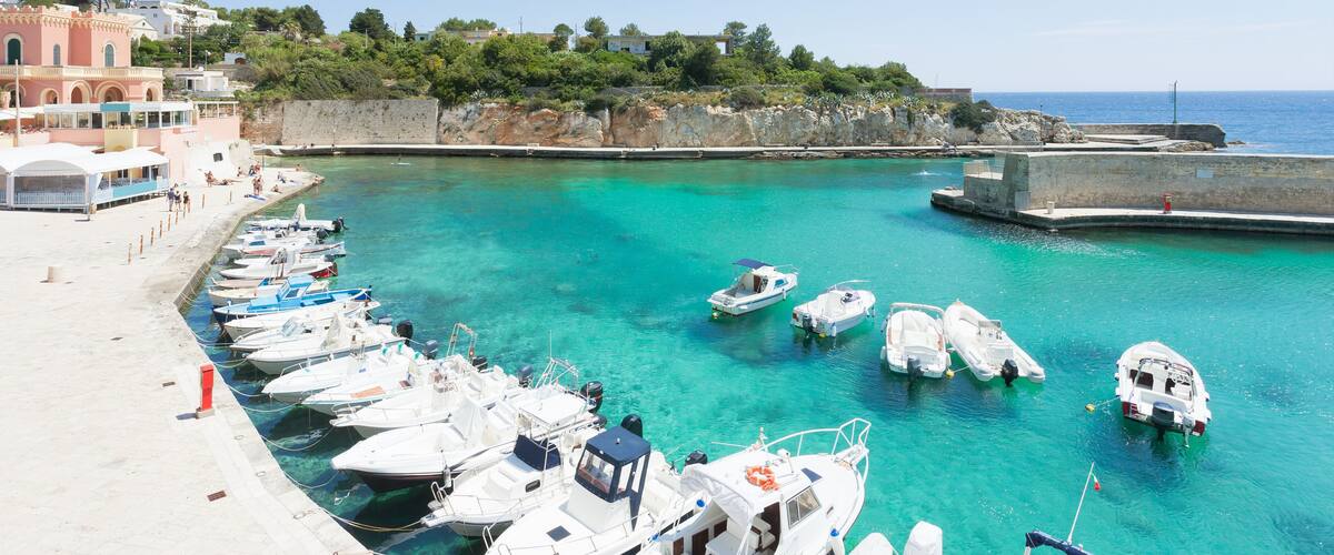 A marina with boats docked along the shore, a clear blue sea, and buildings in the background.