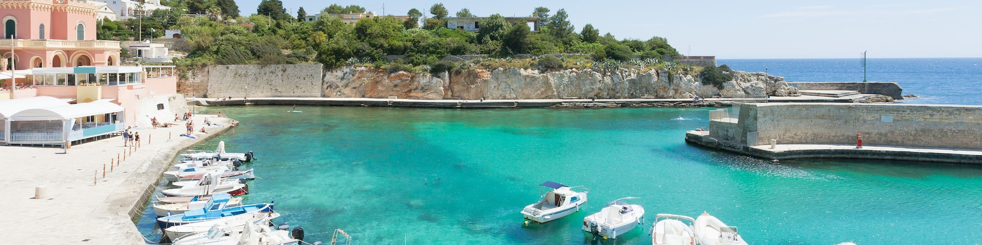 A marina with boats docked along the shore, a clear blue sea, and buildings in the background.