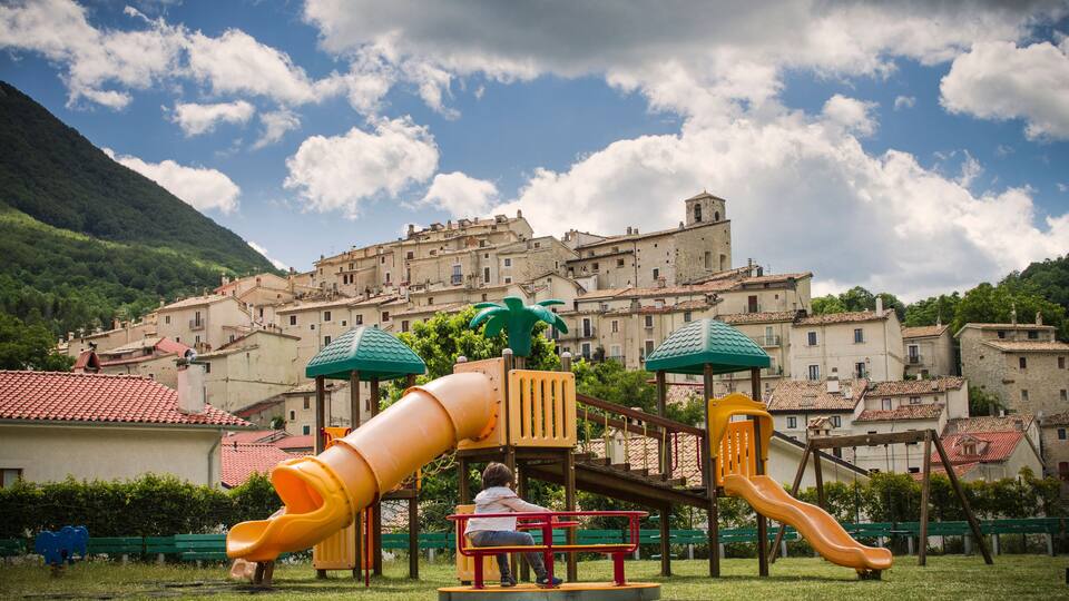 A playground with slides and a child sitting on a bench, set against a backdrop of historic buildings and a mountain.