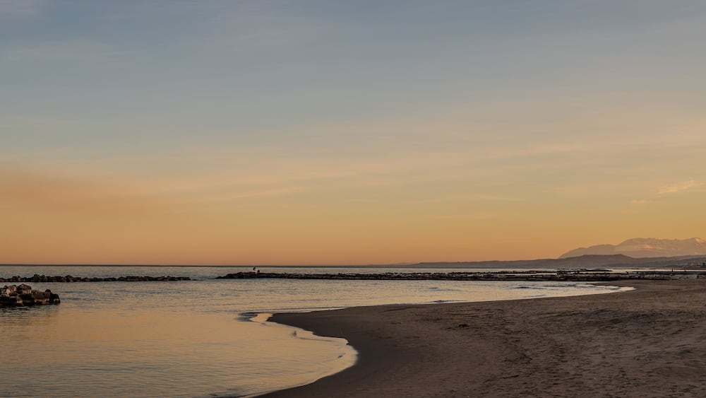 Ein Strand mit einem steinernen Wellenbrecher und einem Berg im Hintergrund.