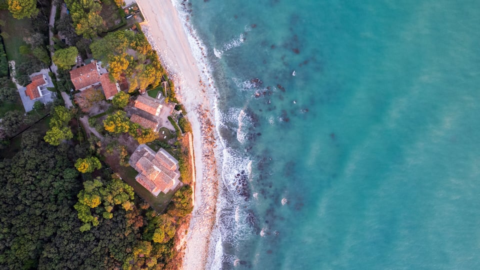 An aerial view of a coastal area with a beach, houses, and dense forest.