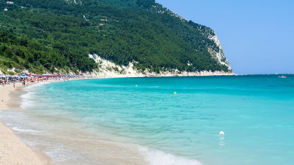 A beach with clear turquoise water, a sandy shore, and a forested hill in the background.