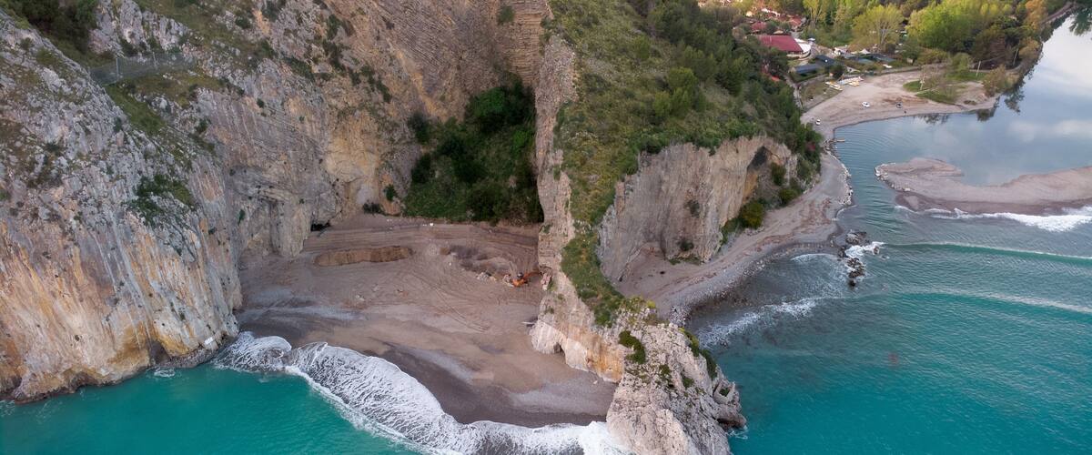 A coastal cliff with a narrow beach, turquoise waters, and a small settlement in the background.