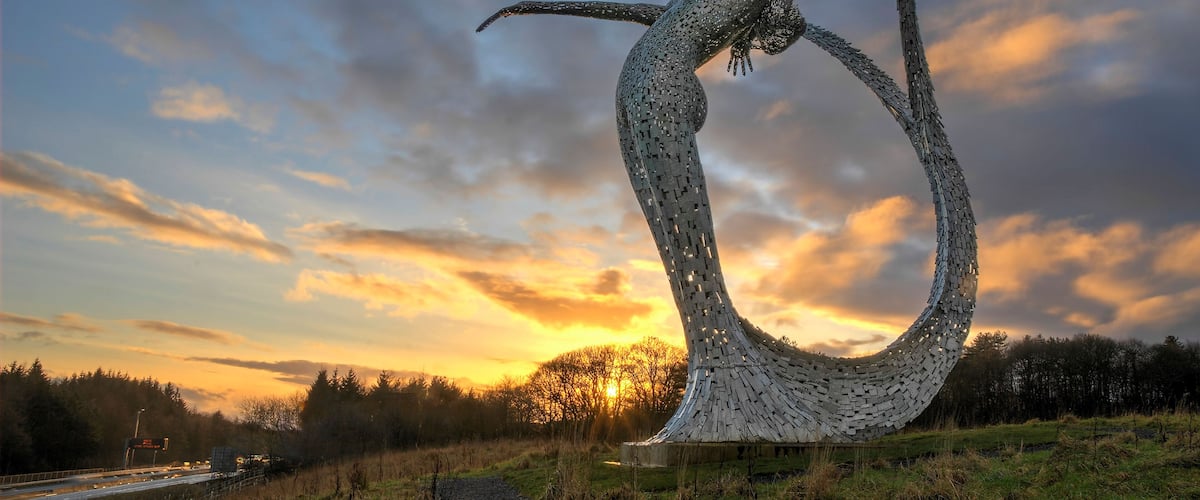 A sculpture of a woman in a dynamic pose, set against a sunset sky with clouds.