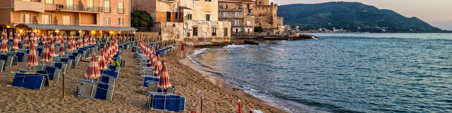 A beach with rows of sunbeds and umbrellas, a red boat, and historic buildings along the coastline.