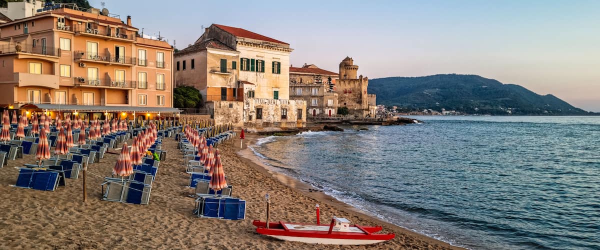 A beach with rows of sunbeds and umbrellas, a red boat, and historic buildings along the coastline.