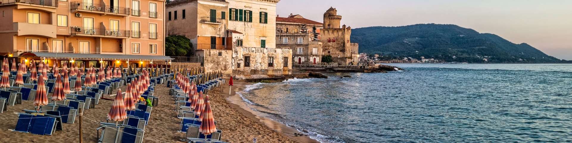 A beach with rows of sunbeds and umbrellas, a red boat, and historic buildings along the coastline.