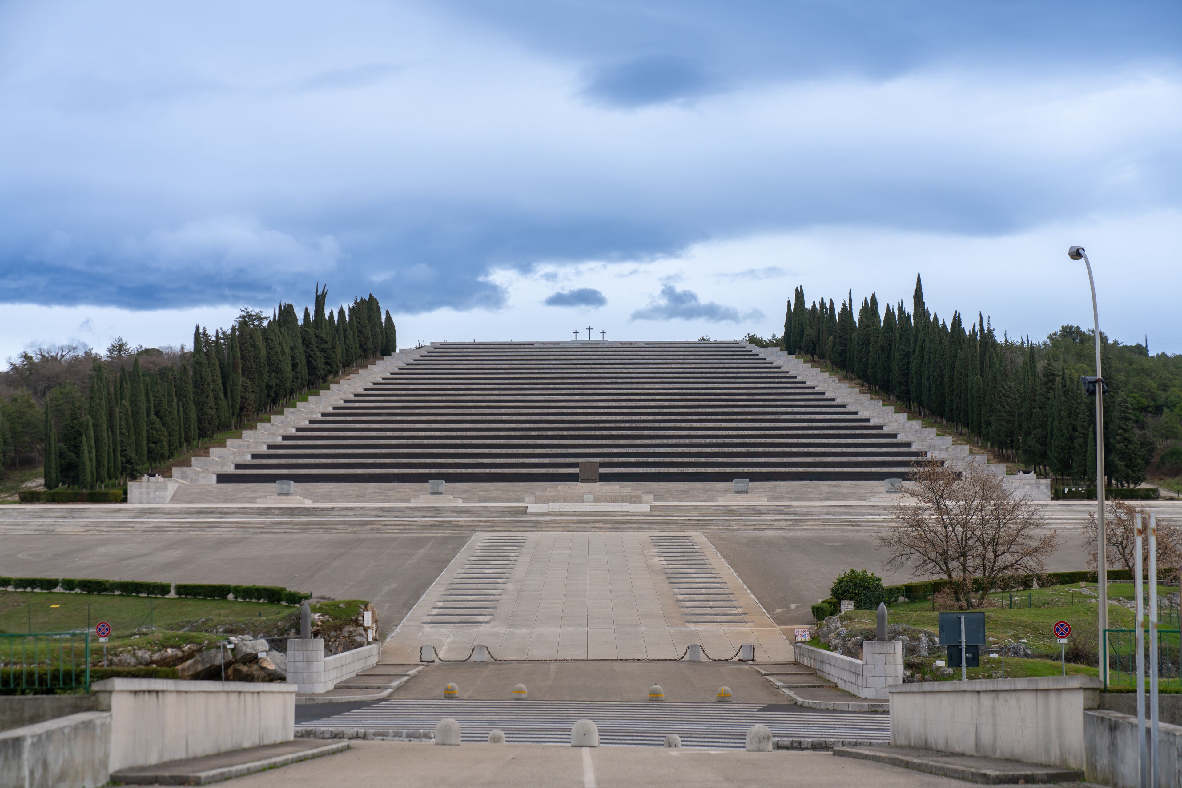 A large, open plaza with a series of steps leading up to a central area, surrounded by trees and a cloudy sky.