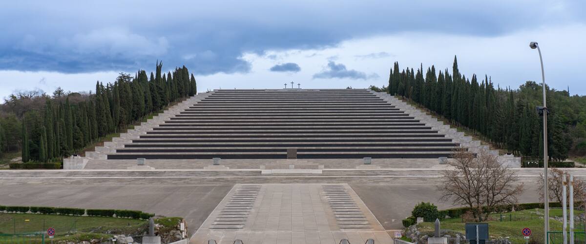 A large, open plaza with a series of steps leading up to a central area, surrounded by trees and a cloudy sky.