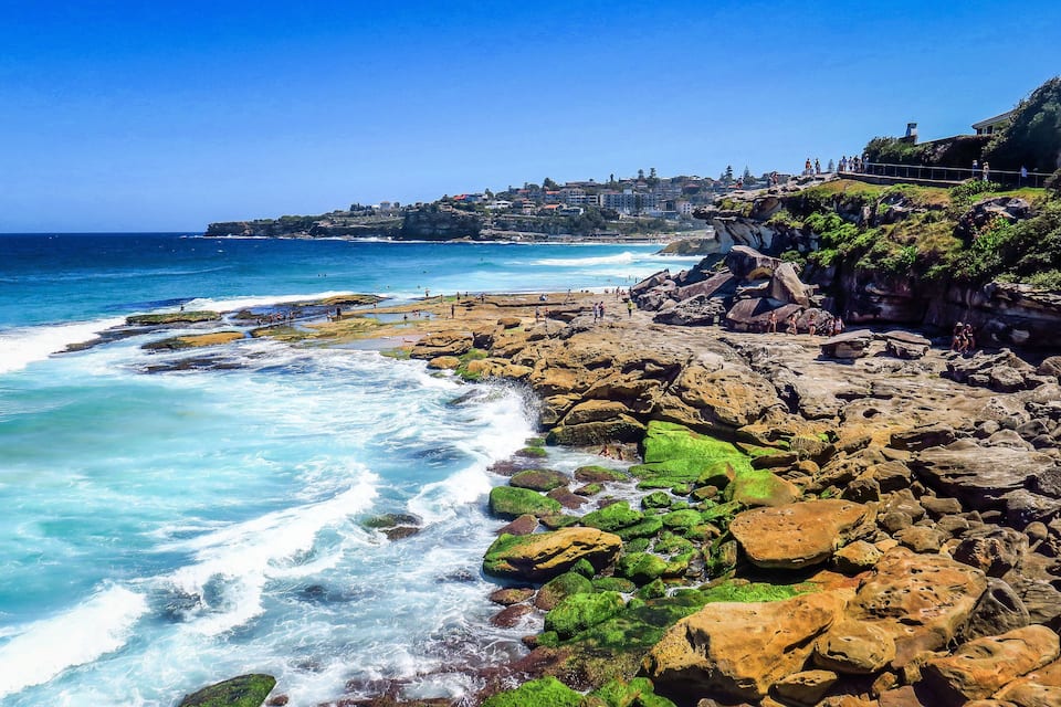 A rocky coastline with waves crashing against the shore, a clear blue sky, and a distant town on a hill.