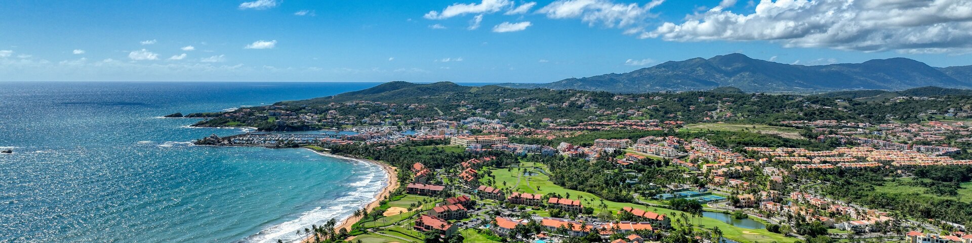 Western Aerial view of Palmas del Mar, Puerto Rico, A southern coast gated community with lush greenery, golf courses, coastline meeting the ocean and rolling hills.