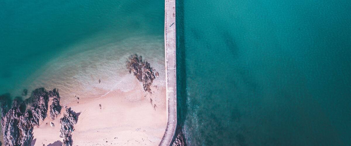 An aerial view of a coastal area with a long pier extending into turquoise waters, a sandy beach, and rocky formations near the shore.