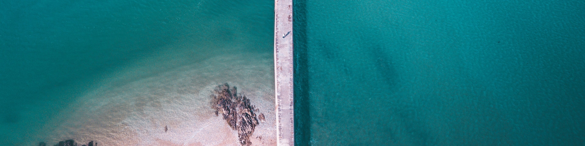 An aerial view of a coastal area with a long pier extending into turquoise waters, a sandy beach, and rocky formations near the shore.