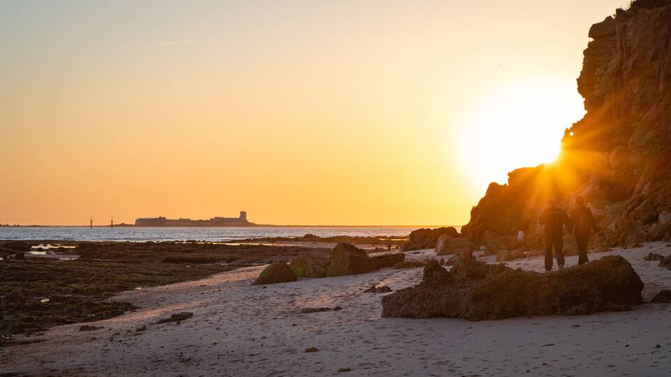 A rocky beach at sunset with the sun setting behind a cliff.