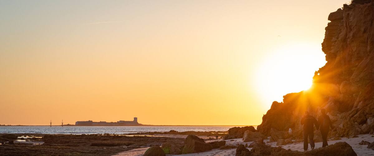 A rocky beach at sunset with the sun setting behind a cliff.
