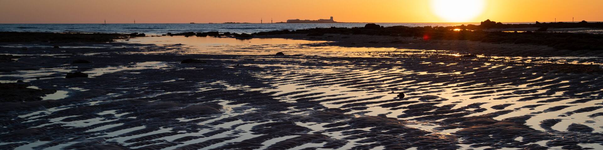 Una playa al atardecer, con el sol bajo en el horizonte, proyectando un resplandor cálido.