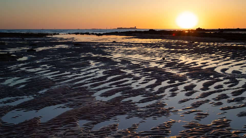 A beach at sunset with the sun low on the horizon, casting a warm glow.