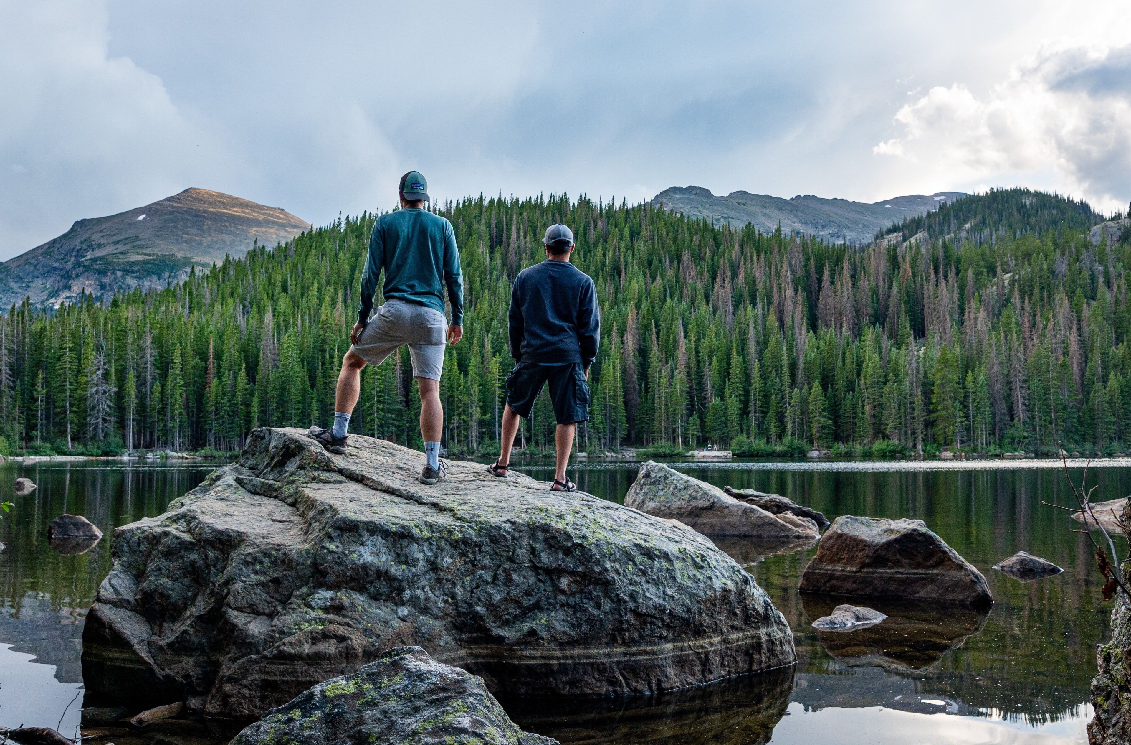 Two people standing on a rock by a lake with mountains in the background.