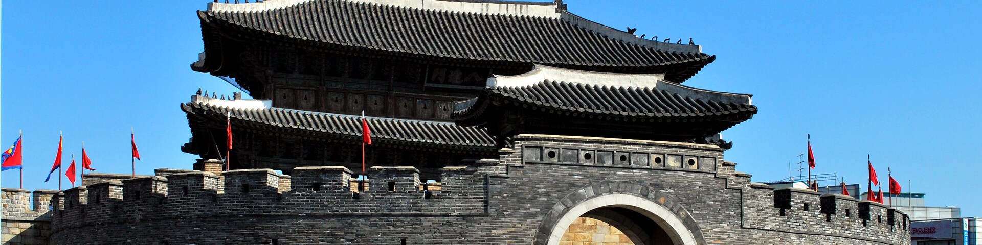 Una puerta tradicional china con varios niveles y banderas rojas, una camioneta blanca al frente y un cielo azul despejado.