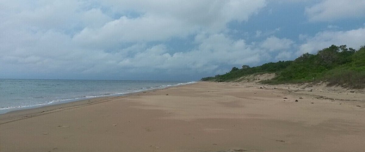 A wide sandy beach with gentle waves, a cloudy sky, and sparse vegetation along the shore.