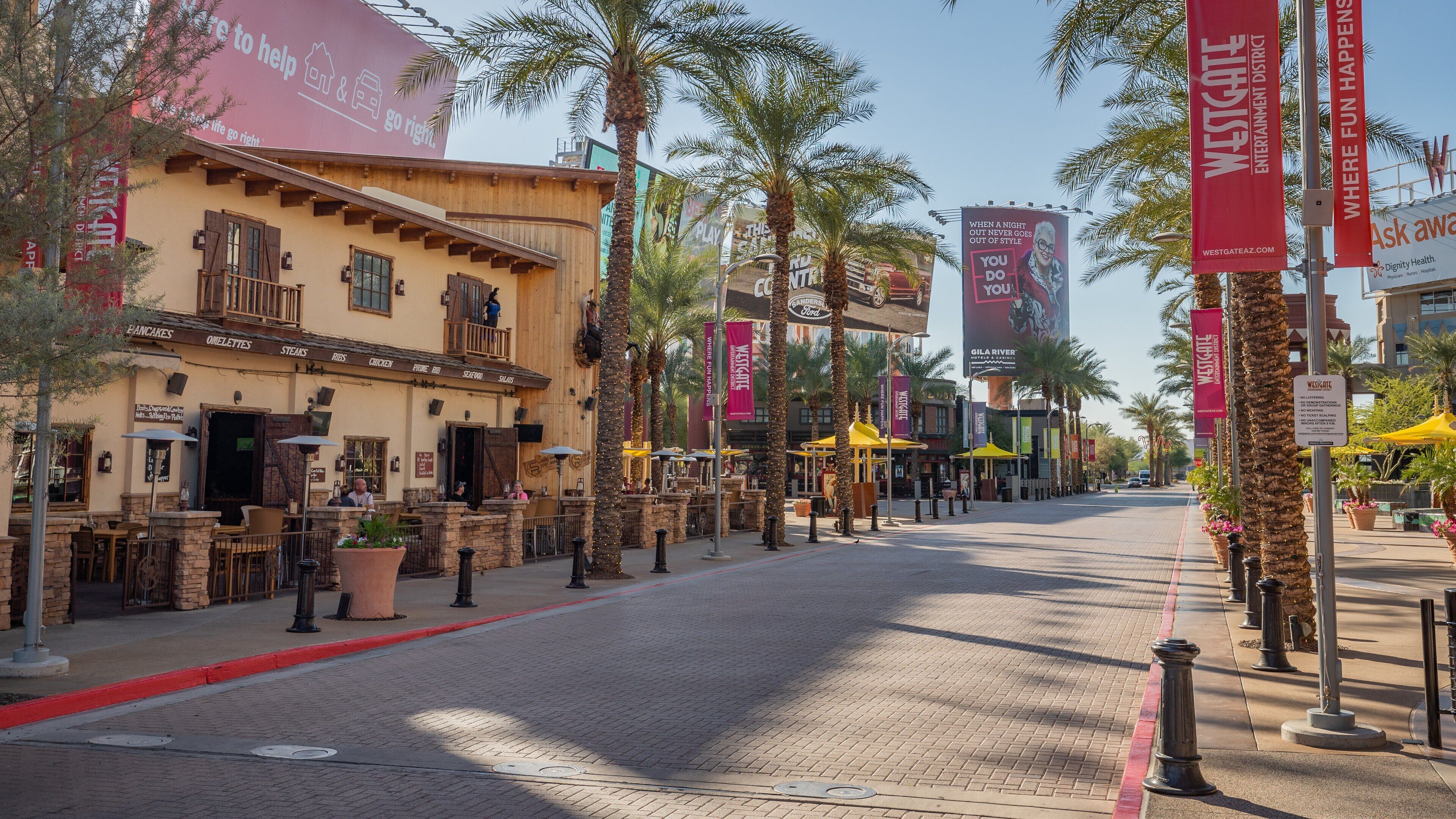Una calle bordeada de palmeras, con terrazas de cafés y pancartas que promocionan comercios locales.