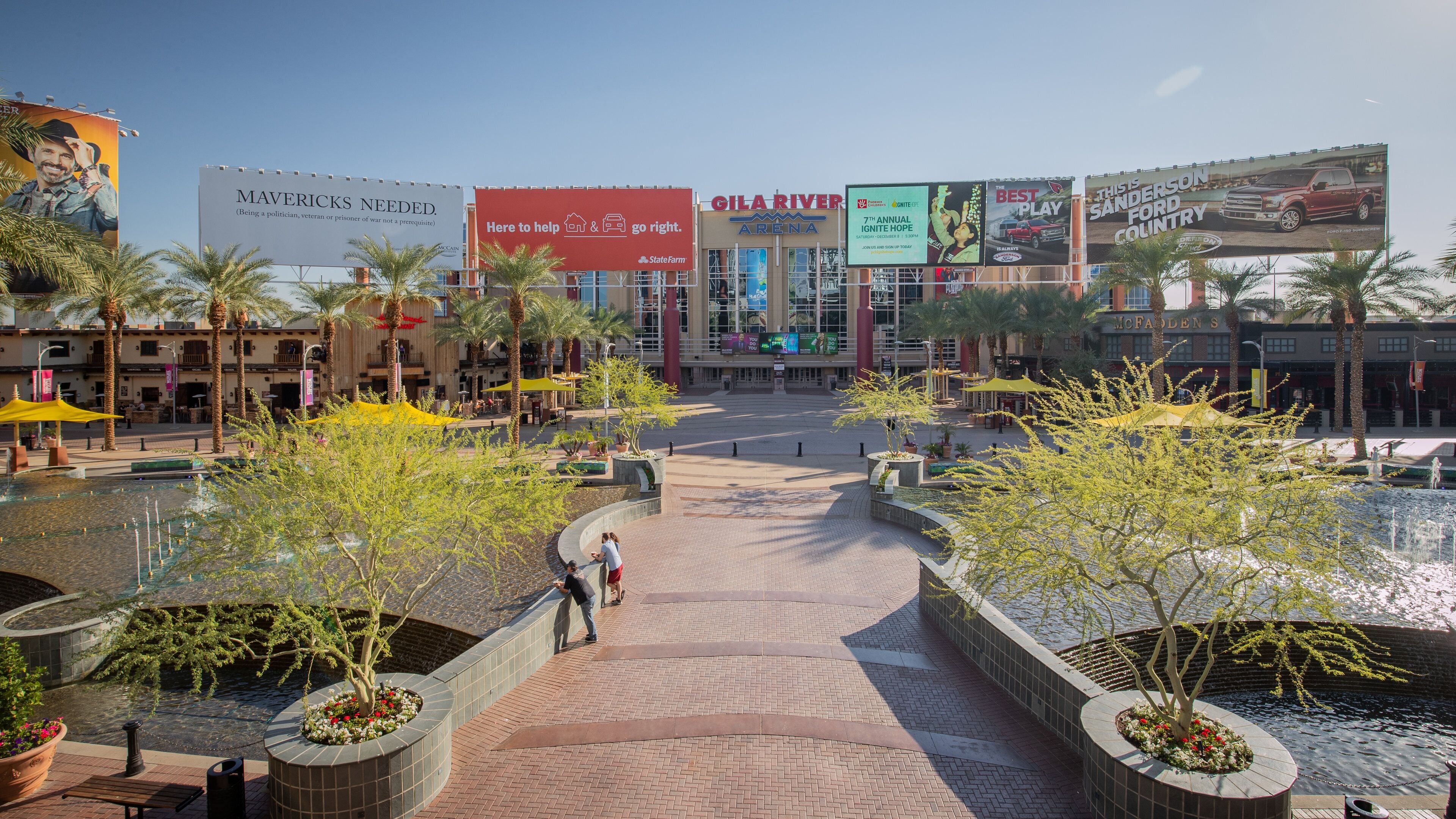 A plaza with palm trees, a fountain, and a large building with advertisements.