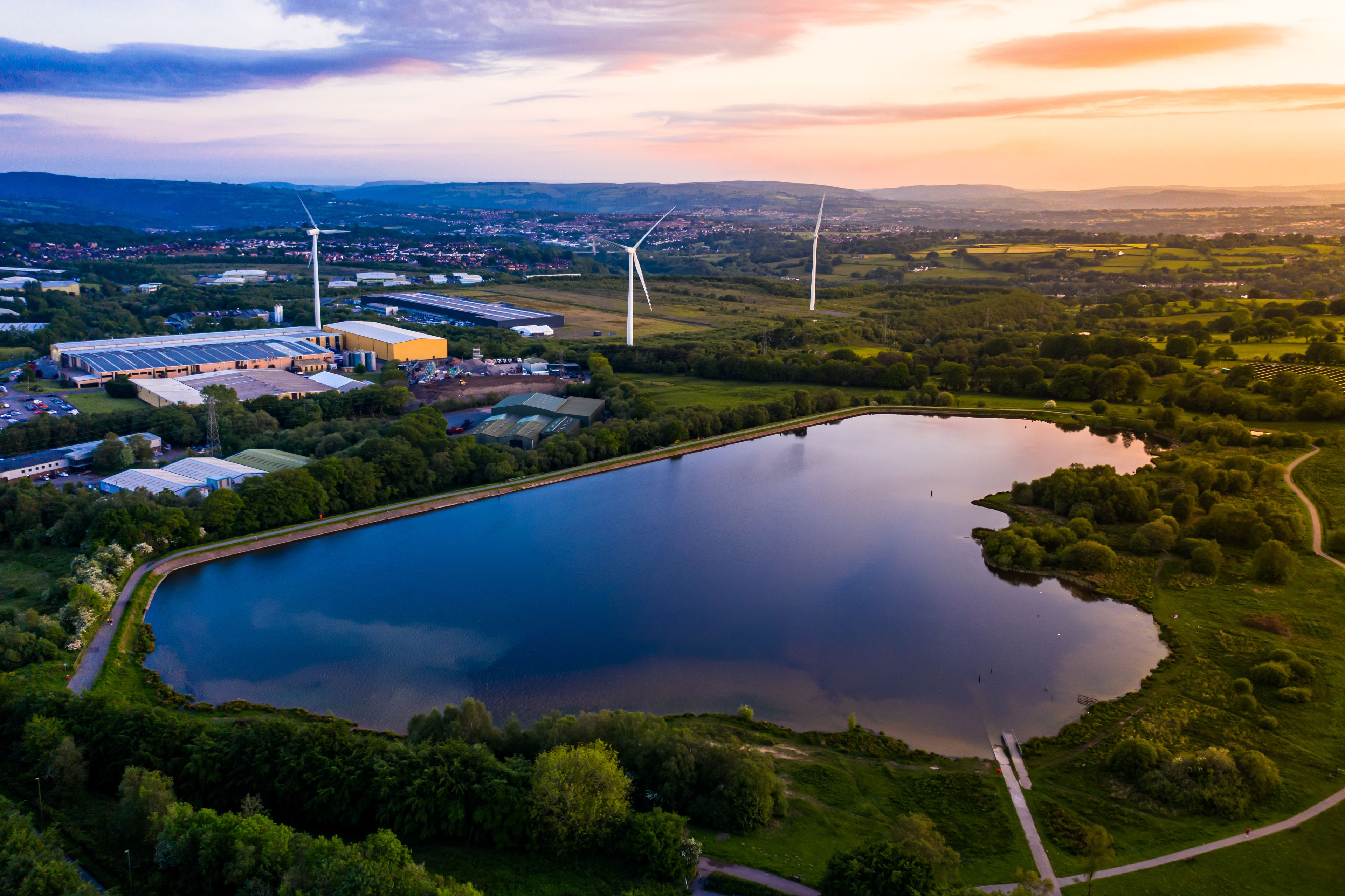 A large body of water surrounded by greenery, with wind turbines and industrial buildings in the background.