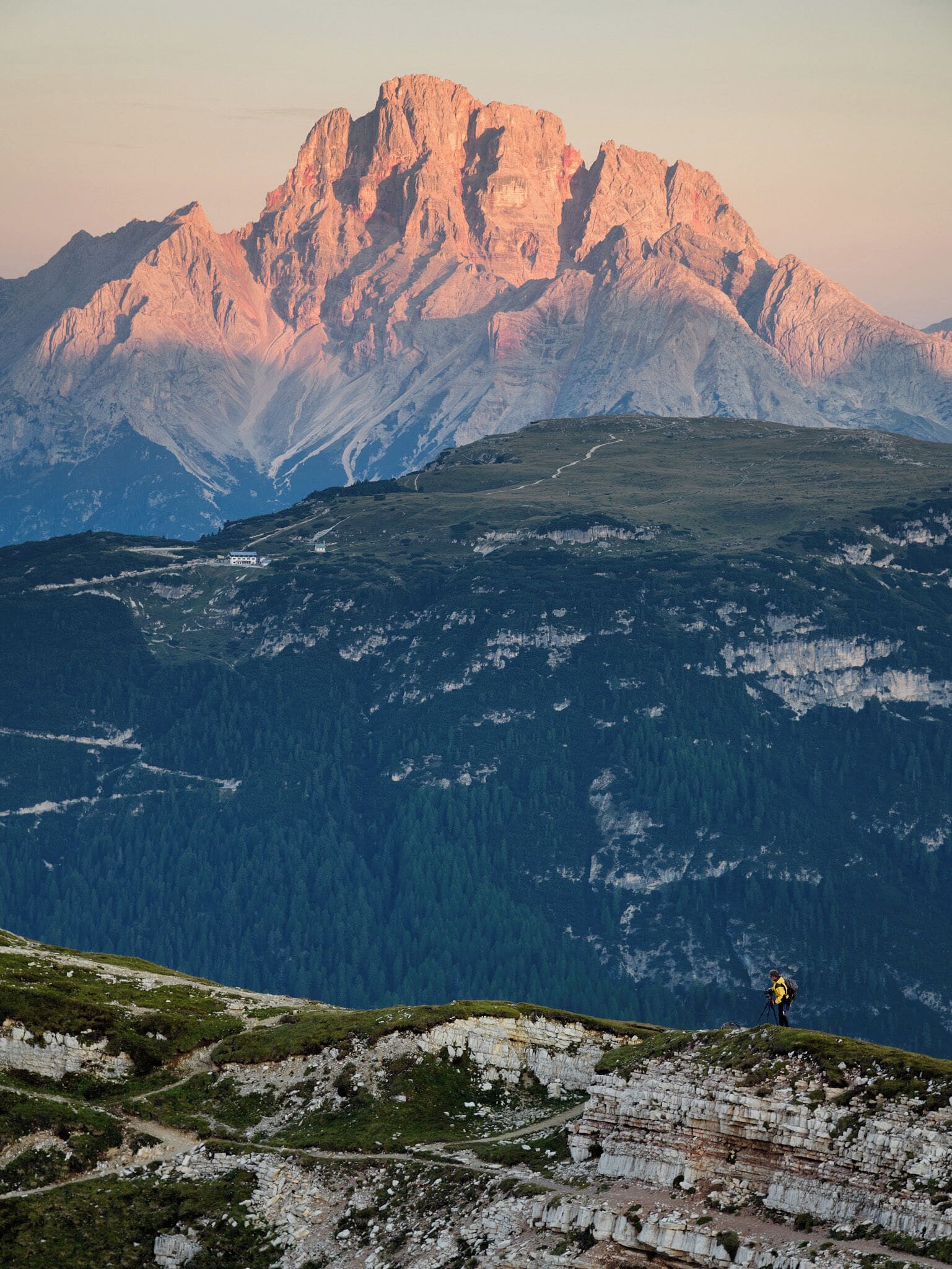 A mountain range with a prominent peak, a hiker with a tripod, and a valley with dense forest.