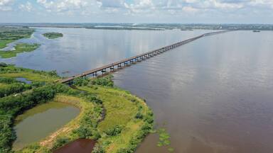 Uma ponte longa sobre um rio largo, com margens verdes e pequenas lagoas.