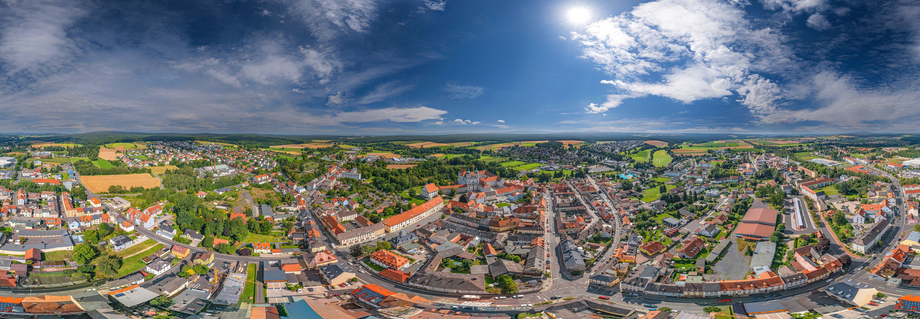A panoramic view of a town with a river, surrounded by fields and hills.
