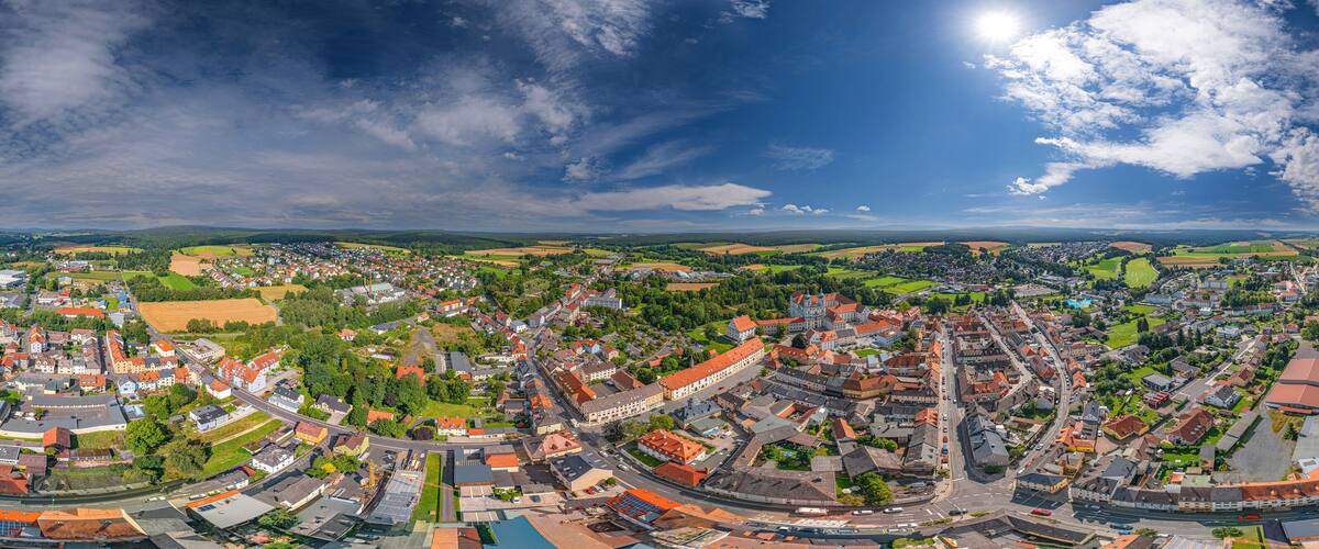 A panoramic view of a town with a river, surrounded by fields and hills.