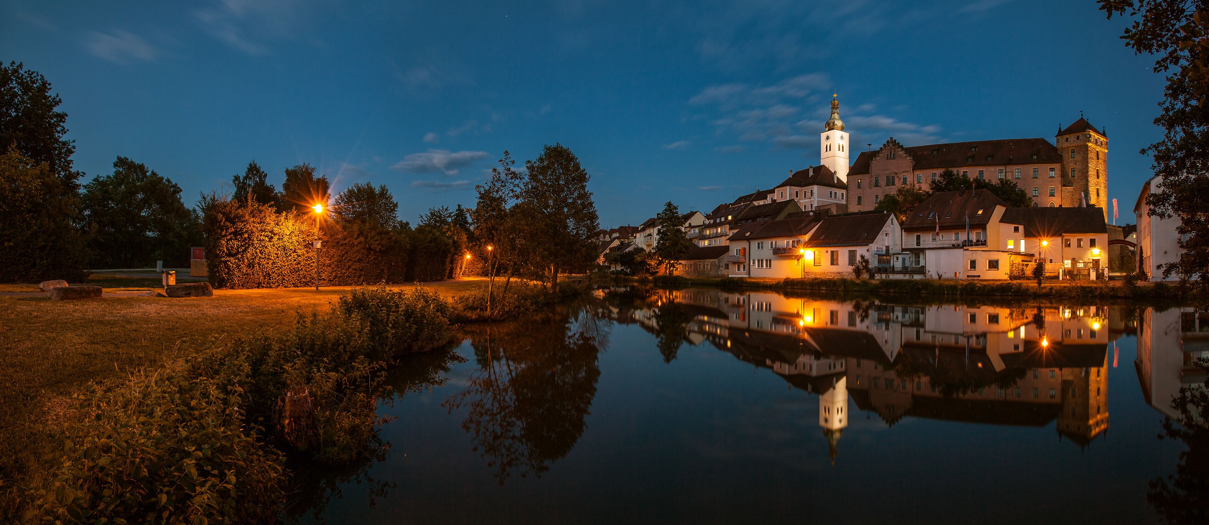 A serene lakeside town at dusk with illuminated buildings reflecting on the water.