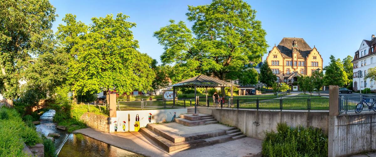 A riverside walkway with steps leading to a gazebo, surrounded by lush greenery and historic buildings.