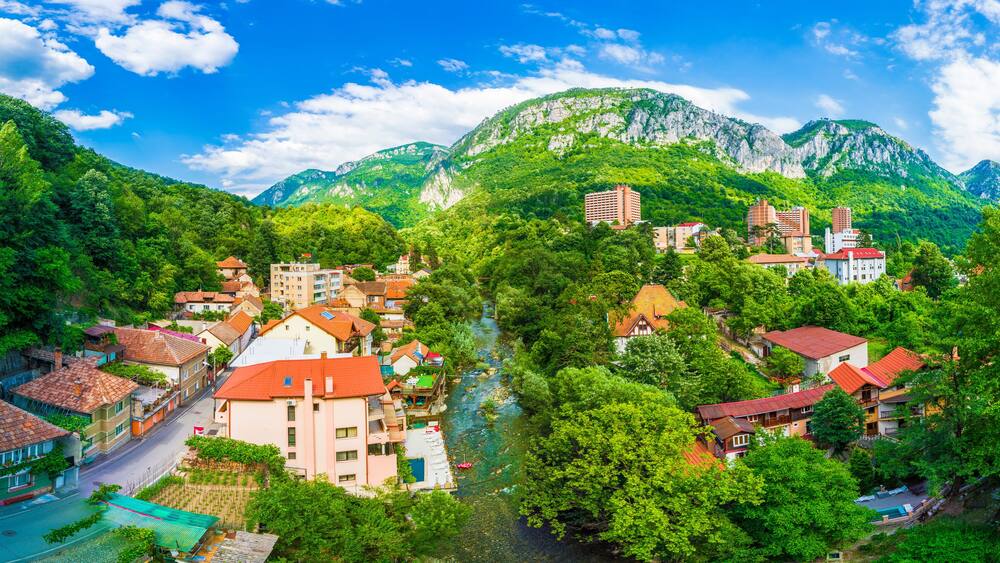 A town nestled in a valley with a river, surrounded by lush green mountains and red-roofed buildings.