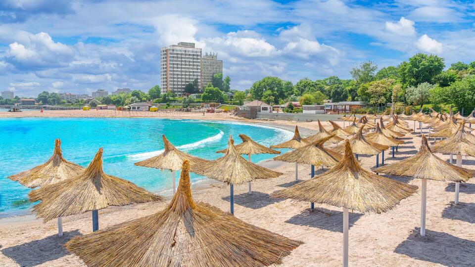 A beach with thatched umbrellas and a building in the background.