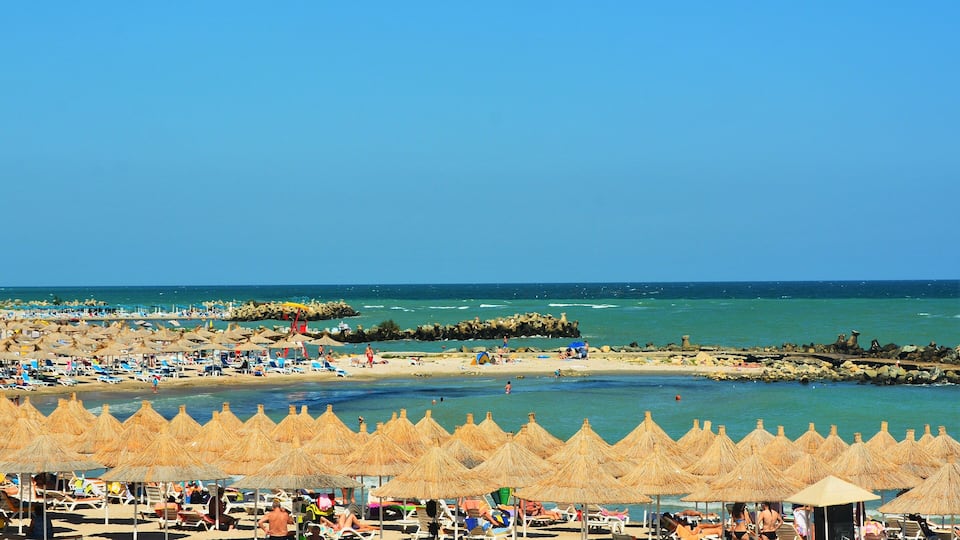 A beach with thatched umbrellas, sun loungers, and a rocky breakwater.