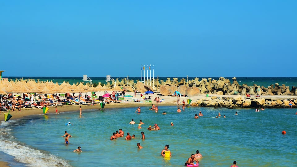 A crowded beach with people swimming and sunbathing, a rocky breakwater, and beach umbrellas.