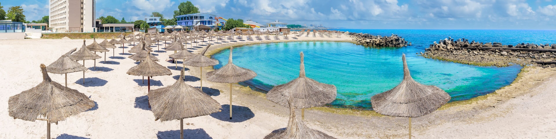 A beach with thatched umbrellas, a pool, and a building in the background.