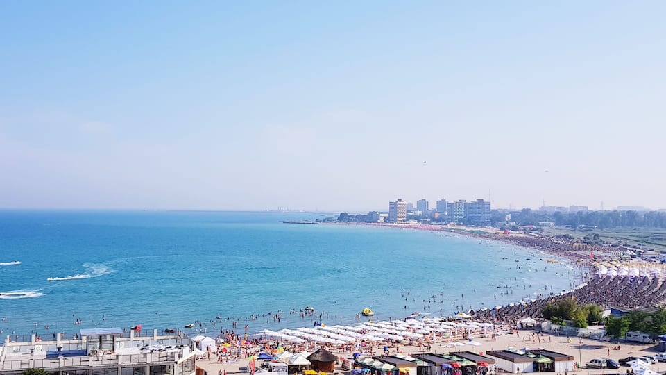 A crowded beach with a long pier extending into the sea, surrounded by buildings.