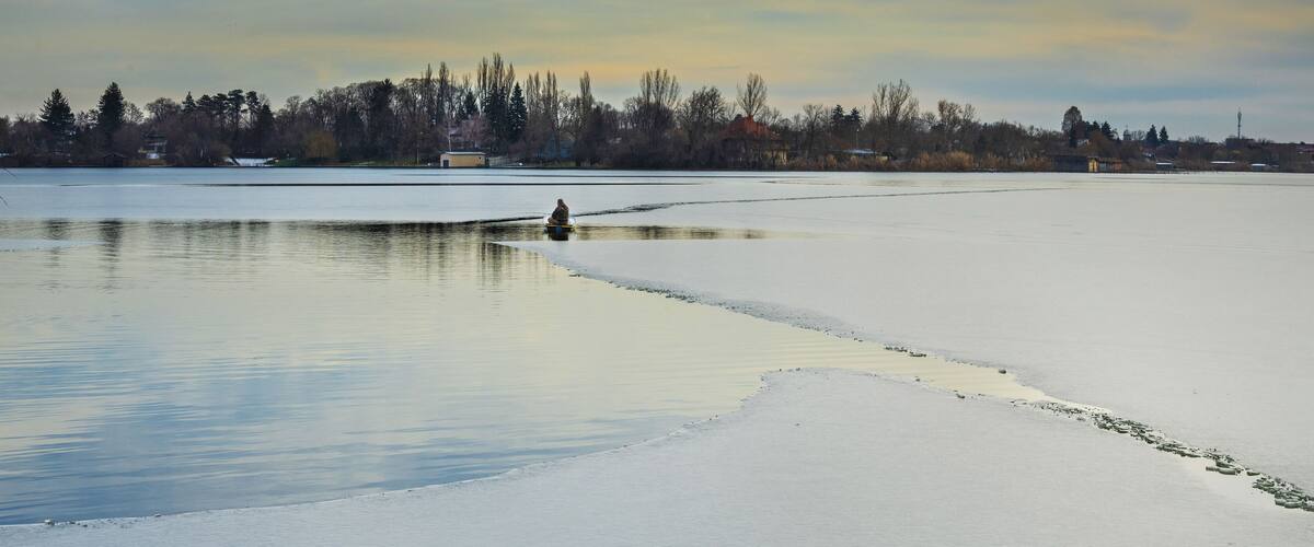 A frozen lake with a person standing in the middle, surrounded by trees and buildings in the background.